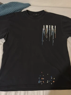 Svart AMIRI t-shirt med färgstänk - Säljer en svart t-shirt från AMIRI med färgstänk och droppande logga i vitt, blått, orange och gult både fram och bak. T-shirten har normal passform och är kortärmad, tillverkad i mjuk bomull. Perfekt för dig som gillar streetwear och unika detaljer.
