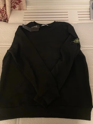 Svart sweatshirt från Stone Island - Svart sweatshirt från Stone Island med klassisk logotyp-patch på vänster ärm. Tröjan har rund halsringning och ribbade muddar vid ärmslut och nederkant. Tillverkad i mjukt bomullsmaterial för en skön och stilren look.
