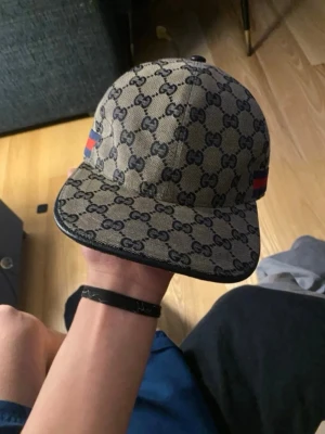 Gucci keps  - Säljer en snygg Gucci keps i beige med klassiskt GG-monogram över hela kepsen. Kepsen har en rak skärm och detaljer med röd och blå rand på sidan. Tillverkad i canvasmaterial och justerbar baktill. Perfekt för dig som vill ha en exklusiv och trendig look.