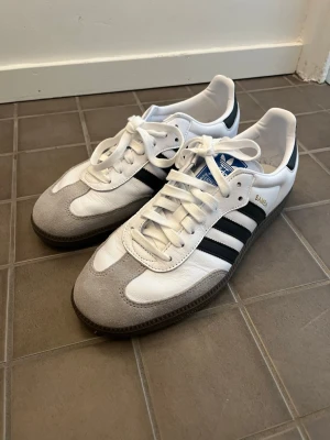 Adidas Samba OG vita och svarta sneakers - Trendiga Adidas Samba OG sneakers i vitt läder med grå mockadetaljer och svarta ränder på sidorna. Endast använda en gång därav priset!!
