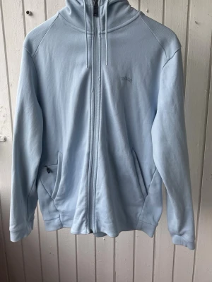 Ljusblå hoodie från BOSS - Ljusblå hoodie från BOSS med dragkedja hela vägen och två fickor framtill, varav en med dragkedja. Diskret BOSS-logga på bröstet. Hoodien har huva och är tillverkad i mjukt material, perfekt för en avslappnad stil. Inga fläckar eller defekter