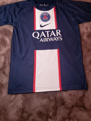 PSG matchtröja blå Nike - Snygg blå PSG matchtröja från Nike med vita och röda detaljer. Tröjan har klubbmärke och Qatar Airways-tryck på bröstet samt korta ärmar. Perfekt för dig som älskar fotboll och vill representera Paris Saint-Germain.