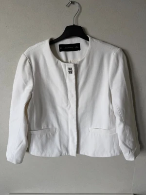 Vit kavaj från Zara Basic - Stilren vit kavaj från Zara Basic med rundad krage och dold knäppning framtill. Kavajen har två fickor. Kavajen har aldrig använts men har tyvärr inte prislappen kvar. 