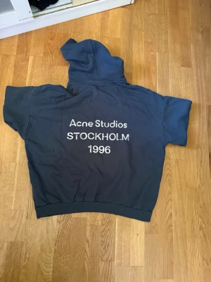 Mörkblå hoodie från Acne Studios - Säljer en mörkblå hoodie från Acne Studios med tryck på ryggen där det står 'Acne Studios STOCKHOLM 1996'. Hoodien har klassisk huva och ribbade muddar. Perfekt för dig som gillar stilrena och trendiga plagg. Priset kan självklart diskuteras! 