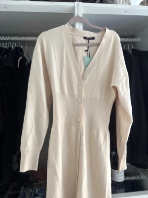 Beige lång stickad klänning - Lång beige stickad klänning med v-ringning och knappar framtill. Klänningen har långa ärmar och mjukt, skönt material