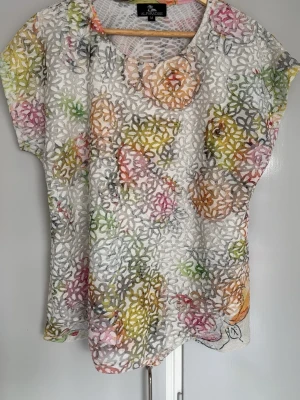 Blommig spetsblus från Al Paradise - Säljer en blommig blus från Al Paradise i vitt med färgglada blommor i rosa, gul, grön och orange. Blusen har rund halsringning, korta ärmar och är gjord i ett tunt, transparent spetsmaterial. Perfekt för dig som gillar unika och luftiga toppar!