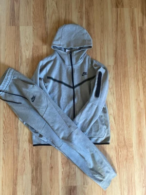 Gråa Nike Tech Fleece mjukisbyxor - Säljer ett par gråa Nike Tech Fleece mjukisbyxor med svarta detaljer och dragkedjor. Byxorna har en slim fit passform och är tillverkade i mjukt och stretchigt material. Perfekta för chill eller träning. Nike-logga på benet och praktiska fickor.