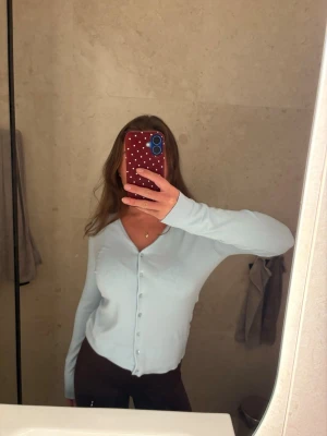 Ljusblå cardigan med v-ringning - Snygg ljusblå cardigan med v-ringning och knappar framtill. Tröjan har långa ärmar och är tillverkad i ett tunt, mjukt material som sitter skönt på kroppen. Perfekt att slänga över en topp eller bära som den är.