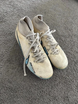 Nike Air Zoom vita fotbollsskor - Säljer ett par vita Nike Air Zoom fotbollsskor med högt skaft och snörning. Skorna har en svart Nike-logga på sidan och detaljer i blått och turkost. Tillverkade i syntetmaterial för bra grepp och komfort på planen. Perfekta för dig som vill ha stilrena och lätta skor.