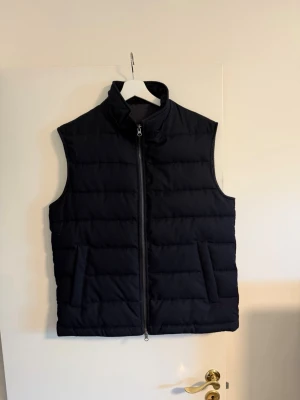 Väst Bruun & Stengade - Stilren mörkblå dunväst med hög krage och hel dragkedja framtill. Västen har två snedställda fickor och quiltad design med horisontella sömmar. Perfekt för lager-på-lager och enkel att matcha till olika outfits.
