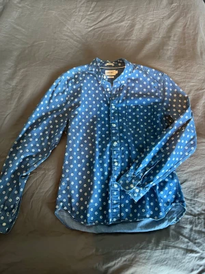 Blå skjorta från East West - Säljer en blå skjorta från East West i slim fit med vita blommor över hela tyget. Skjortan har långa ärmar, knappar framtill och en klassisk krage. Perfekt för dig som gillar en unik och fräsch stil under sommaren. Mindre passform, skulle kladda den som M.