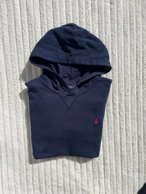 Mörkblå hoodie från Polo Ralph Lauren - Snygg mörkblå hoodie från Polo Ralph Lauren med klassisk huva och känguruficka. Ikoniska röda loggan broderad på bröstet. Tillverkad i mjuk bomull som känns skön mot huden. Perfekt för en chill och stilren look.