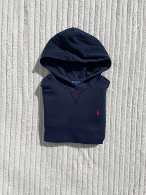 Mörkblå hoodie från Polo Ralph Lauren - Mörkblå hoodie från Polo Ralph Lauren med klassisk huva och känguruficka. Tröjan har en liten röd broderad logga på bröstet och är gjord i mjukt bomullsmaterial. Perfekt för en avslappnad och stilren look.