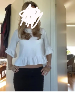 Vit volangblus med trekvartsärm - Söt vit blus med volangkant nedtill och trekvartsärm. Toppen har en croppad passform och är perfekt för dig som gillar en feminin och trendig stil. Materialet känns mjukt och lätt, och toppen har en enkel men chic look.