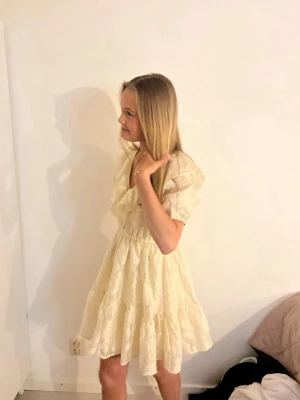 Ljus kort volangklänning med spets - Supersöt ljus kort klänning med volanger och v-ringning. Klänningen har puffärmar, markerad midja och är dekorerad med spetsdetaljer. Perfekt för sommarens alla tillfällen!