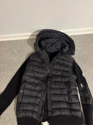 Svart pufferjacka från Moncler - Säljer en svart pufferjacka från Moncler med quiltad front och huva. Jackan har stickade ärmar med dragkedjedetalj och Moncler-logga på ena ärmen. Perfekt för kalla vinterdagar och riktigt snygg streetstil.