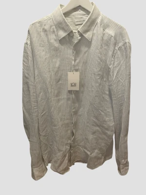 Randig linneskjorta från These Glory Days - Snygg vit skjorta med tunna blå ränder från These Glory Days. Skjortan är i slim fit-modell och tillverkad i 100% linne, perfekt för en stilren och luftig look. Klassisk krage och långa ärmar med knäppning framtill.