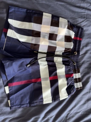 Burberry shorts  - Snygga badbyxor i marinblått med klassiskt rutmönster i vitt, brunt och svart samt röda detaljer. De har elastisk midja med snörning och en bakficka. Perfekta för stranden eller poolen och tillverkade i snabbtorkande material.