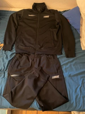 Svart Nike träningsset herr - Säljer ett svart träningsset från Nike med zip-jacka och matchande byxor. Setet har vita Nike-loggor på både jacka och byxa samt dragkedja framtill på jackan. Materialet är mjukt och stretchigt, perfekt för träning eller chill. Byxorna har resår i midjan.