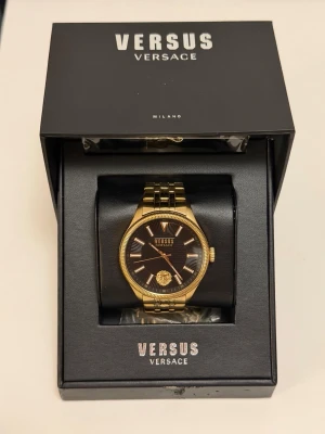 Versus Versace Colonne Gold/Black – Herrklocka – Komplett med box & extra länkar - Versus Versace Colonne herrklocka i guldfärgat rostfritt stål med svart urtavla och ikoniskt lejonmotiv vid kl. 6.  VSPHI0620 (Colonne-serien).  Klockan är i fint begagnat skick och fungerar som den ska. Kommer med:  * Originalbox * Manual/häfte * Extra länkbitar för justering * Originalförpackning  Specifikationer:  * Modell: Versus Versace Colonne * Referens: VSPHI0620 * Quartzverk (batteridriven) * Guldfärgat rostfritt stål * Mineralglas * Vattentålig 30 m / 3 ATM * Diameter ca 45 mm