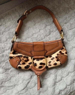 Brun dolce & ga handväska med leopardmönster - Säljer en unik handväska i brunt läder med leopardmönstrad pälsdetalj. Väskan har guldfärgade metalldetaljer, justerbart axelband och coola nitar på sidorna. Perfekt accessoar för dig som vill sticka ut med en trendig och djurinspirerad look. Små spår av användning