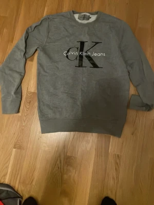 Grå sweatshirt från Calvin Klein Jeans - Snygg grå sweatshirt från Calvin Klein Jeans med stort svart CK-tryck och vit logga på bröstet. Tröjan har rund halsringning, långa ärmar och ribbade muddar. Perfekt för en avslappnad och trendig look.