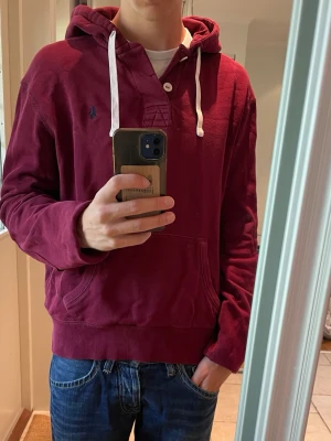 Vinröd hoodie från Polo Ralph Lauren - Snygg vinröd hoodie från Polo Ralph Lauren med vit dragsko i huvan och känguruficka framtill. Klassisk logga broderad på bröstet och ribbade muddar vid ärmar och nederkant. Perfekt för en avslappnad och stilren look.