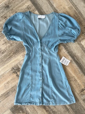 Ljusblå kort jeansklänning med puffärm - Supersöt ljusblå kort klänning i mjukt jeansmaterial med puffärmar och v-ringning, från Asos. Klänningen har knappar framtill och markerad midja för en snygg siluett. Klänningen är i nytt skick med lappen kvar, den har en liten defekt (se bild 6 och 7, och bredvid knapp 2) i form av ett litet trådsläpp, men inget som syns när den är knäppt eller från insidan eller som påverkar användningen! Hör av er vid funderingar!😊