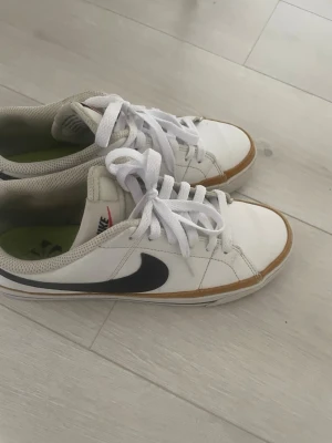 Vita Nike sneakers med svart swoosh - Snygga vita Nike sneakers med svart swoosh på sidan och beige/brun detalj runt sulan. Klassisk låg modell med vita skosnören och meshfoder. Perfekta för en avslappnad streetwear-look.