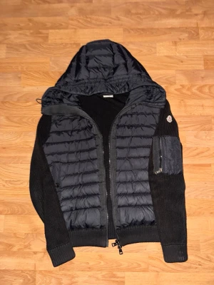 Svart pufferjacka med huva från Moncler - Snygg svart pufferjacka från Moncler med stickade ärmar och vadderad kropp. Jackan har huva, dragkedja framtill och en ficka med dragkedja på ärmen. Perfekt för kyliga dagar och ger en sportig look.