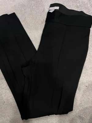 Svarta leggingsbyxor med hög midja från H&M - Snygga svarta leggingsbyxor med hög midja och raka ben. Byxorna har en stilren design med pressveck och en dekorativ bakficka. Tillverkade i ett stretchigt material som ger en bekväm passform. Perfekta för en clean och trendig look.