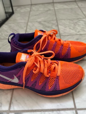 Nike Flyknit Lunar 2 orange/lila 36,5 - Nike Flyknit Lunar 2 sneakers i en färgstark kombination av orange och lila. Ovandelen är i ventilerande mesh med stickade detaljer och vita Nike-loggor på sidan. Skorna har låga skaft och en lätt, dämpad sula för extra komfort. Perfekta för dig som gillar sportig stil. Nike Flyknit Lunar 2 Atomic Orange Dam Sneakers Vit-Court-Lila-Röd-Violett