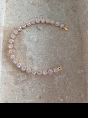 Guldigt armband med gnistrande stenar - Snyggt armband i guldton med runda, gnistrande stenar i hela länken. Armbandet har klassisk tennisdesign och stängs med ett smidigt lås. Perfekt för dig som gillar stilrena och glittriga accessoarer.
