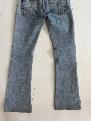 Blå bootcut jeans från G-Rise Gena Jeans - Säljer ett par ljusblå bootcut jeans från G-Rise Gena Jeans med broderade stjärnor på bakfickorna. Klassisk femficksmodell med snygga slitningar och hög midja. Perfekta för dig som gillar retrovibbar och vill ha ett par unika jeans i garderoben. Längd för en person 155-160 cm. Ursprungspris: 2200kr. 