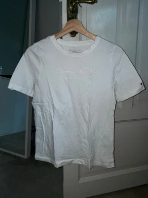 Vit t-shirt från Tommy Hilfiger - Klassisk vit t-shirt från Tommy Hilfiger med diskret broderad logga på bröstet. T-shirten har rund halsringning och korta ärmar. Tillverkad i mjuk bomull som känns skön mot huden. Perfekt basplagg för en clean och enkel stil. Använd fåtal gånger.💕