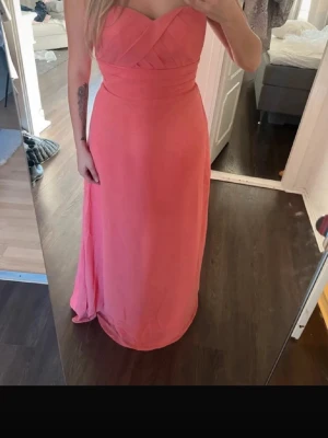 Lång rosa axelbandslös klänning - Säljer en elegant långklänning i rosa med axelbandslös design och omlott-detaljer upptill. Klänningen har en markerad midja och faller snyggt längs kroppen. Perfekt för bal eller festliga tillfällen.