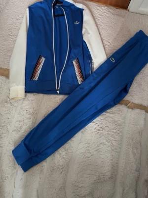 Blå träningsdress från Lacoste Sport - Snygg blå träningsdress från Lacoste Sport med vita ärmar och detaljerade fickor. Setet består av zip-jacka och matchande byxor, båda med Lacoste-logga. Tillverkad i ett mjukt och stretchigt material som passar perfekt till träning eller chill. Använd 1-2 gånger Skick 10/10!!  Koftan storlek XS Byxorna S