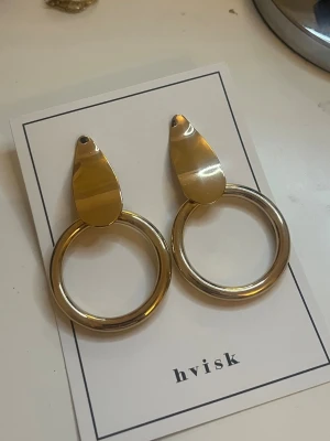 Guldiga statementörhängen från Hvisk - Säljer ett par stora, guldiga statementörhängen från Hvisk. Örhängena har en droppformad platta upptill och en rund, tjock ring nedtill. De är tillverkade i metall med blank finish och har en modern, trendig look.