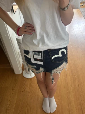 Evisu jeanskjol - Säljer en mörkblå jeanskjol från Evisu med slitna kanter och färgglada loggor på fickorna. Passar mig som brukar ha M/L i byxor. Orginal pris 305$=ca2900kr