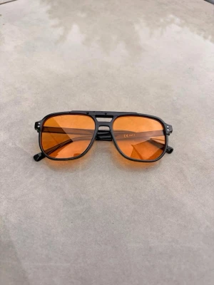 Svarta solglasögon med orange glas - Säljer ett par riktigt snygga solglasögon med svart båge och orangefärgade glas. De har en klassisk pilotform och ger en cool retrovibe. Perfekta för dig som vill sticka ut och addera färg till din stil.🕶️⛱️❤️☀️