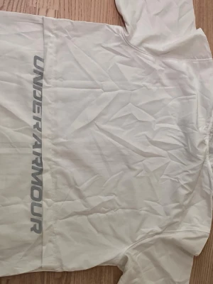 Vit vindjacka från Under Armour - Snygg vit vindjacka från Under Armour med hel dragkedja framtill och diskret logga på bröstet. Jackan har långa ärmar, huva och stora fickor. Materialet är lätt och passar perfekt för blåsiga dagar. Stor Under Armour-tryck längs ryggen.