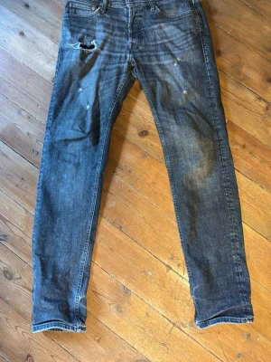 Grå/svart slitna jeans med hål - Säljer ett par blå jeans med skinny fit och slitningar, inklusive ett större hål på vänster lår. Jeansen har klassisk femficksdesign och är tillverkade i denim. Perfekta för en avslappnad och trendig look.