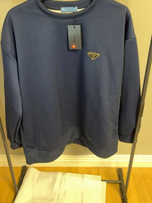 Mörkblå sweatshirt från Prada - Säljer en mörkblå sweatshirt från Prada med klassisk rund halsringning och ribbade muddar. Tröjan har det ikoniska Prada-emblemet i metall på bröstet och är tillverkad i mjuk bomullsblandning. Perfekt för en clean och stilren look.