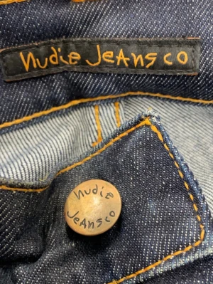 Mörkblå jeans från Nudie Jeans - Snygga mörkblå jeans från Nudie Jeans med klassisk femficksmodell och kontrastsömmar i orange. Jeansen har en rak passform och är tillverkade i slitstark denim av bomull. Perfekta för dig som gillar stilrena och tidlösa jeans.