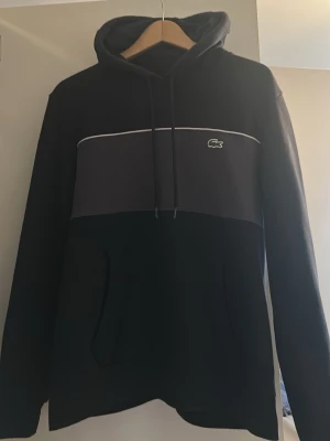 Svart och mörkgrå hoodie från Lacoste - Snygg hoodie från Lacoste i svart och mörkgrått med vit piping över bröstet och den klassiska krokodilloggan. Hoodien har huva med dragsko och en stor magficka. Perfekt för en avslappnad och trendig stil.