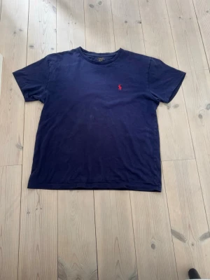 Marinblå t-shirt från Polo Ralph Lauren - Klassisk marinblå t-shirt från Polo Ralph Lauren med röd broderad logga på bröstet. T-shirten har rund halsringning och korta ärmar. Perfekt basplagg för en avslappnad stil.