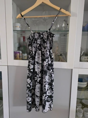 Svartvit blommig sommarklänning - Säljer en svartvit kort klänning med blommigt mönster och smala axelband. Klänningen har en smockad överdel och är i luftig modell, perfekt för varma dagar. Materialet känns lätt och svalt, troligen bomull eller polyester. Passar dig som gillar en avslappnad och feminin stil. Köpt i Sydafrika, oklart märke
