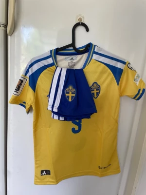 Svensk landslagströja Isak #9 Adidas - Gul och blå svensk landslagströja från Adidas med Isak och nummer 9 på ryggen. Tröjan har korta ärmar, broderade landslagsemblem, Aeroready-material och detaljer i vitt och blått. Matchande blå shorts medföljer. Perfekt för fotbollsfans!