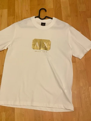 Vit t-shirt från Armani Exchange - Snygg vit t-shirt från Armani Exchange med ett stort guldfärgat logotryck på bröstet. Klassisk rund halsringning och korta ärmar. Tillverkad i mjuk bomull för en bekväm känsla. Perfekt för dig som gillar stilrena plagg med exklusiv touch.
