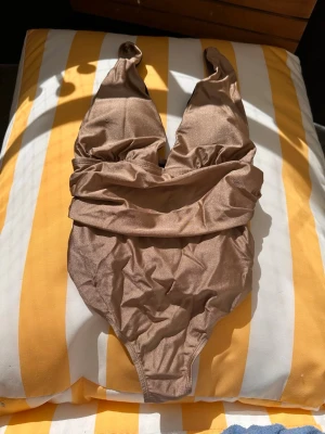 Guldig baddräkt med djup urringning 36/38 - Snygg baddräkt i skimrande bronsfärg med djup urringning och breda axelband. Materialet ser ut att vara stretchigt och följsamt, perfekt för strand eller pool. Draperad detalj över magen ger en cool och trendig look .Storlek 36/38